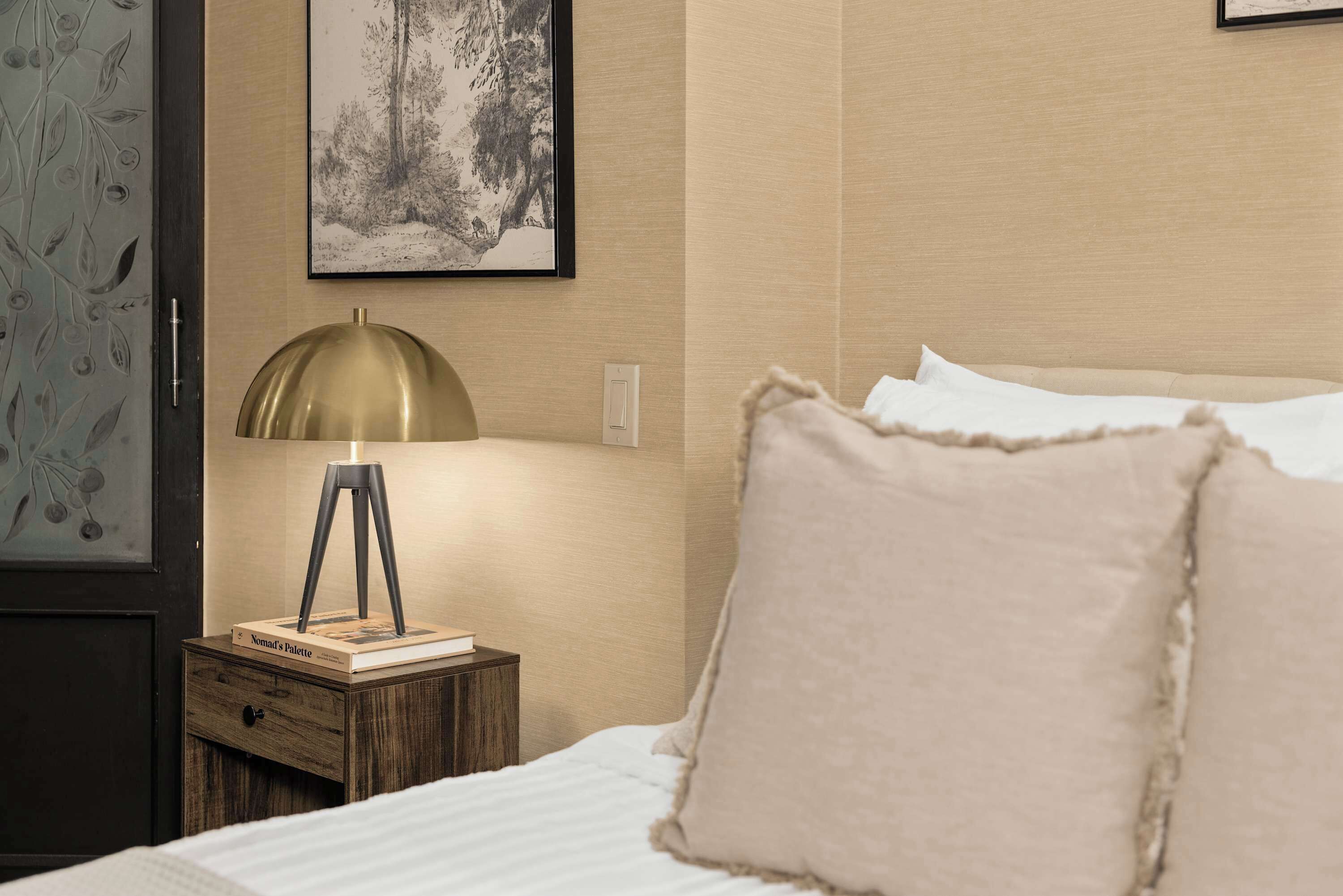 Een keurig opgemaakt bed met een witte dekbedovertrek en beige kussens, een klein houten nachtkastje met een messing lamp, een lijst met een foto aan de muur, en een deur met een ruitpaneel met bladmotief.