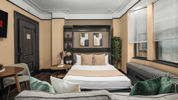 Une chambre d’hôtel avec un lit, un canapé, une petite table, une plante et une fenêtre avec des stores.