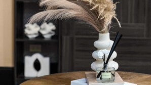 Une table ronde en bois sur laquelle se trouve un vase blanc contenant des plumes d’herbe des pampas, un livre intitulé « BOHEMIA » et un petit flacon de parfum.