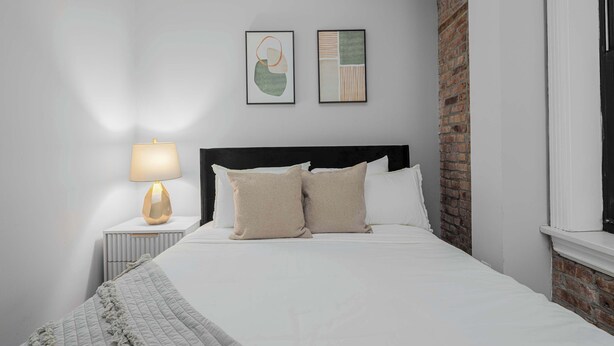 Una cama bien hecha con cabecera, una lámpara de noche y dos cuadros enmarcados en la pared.