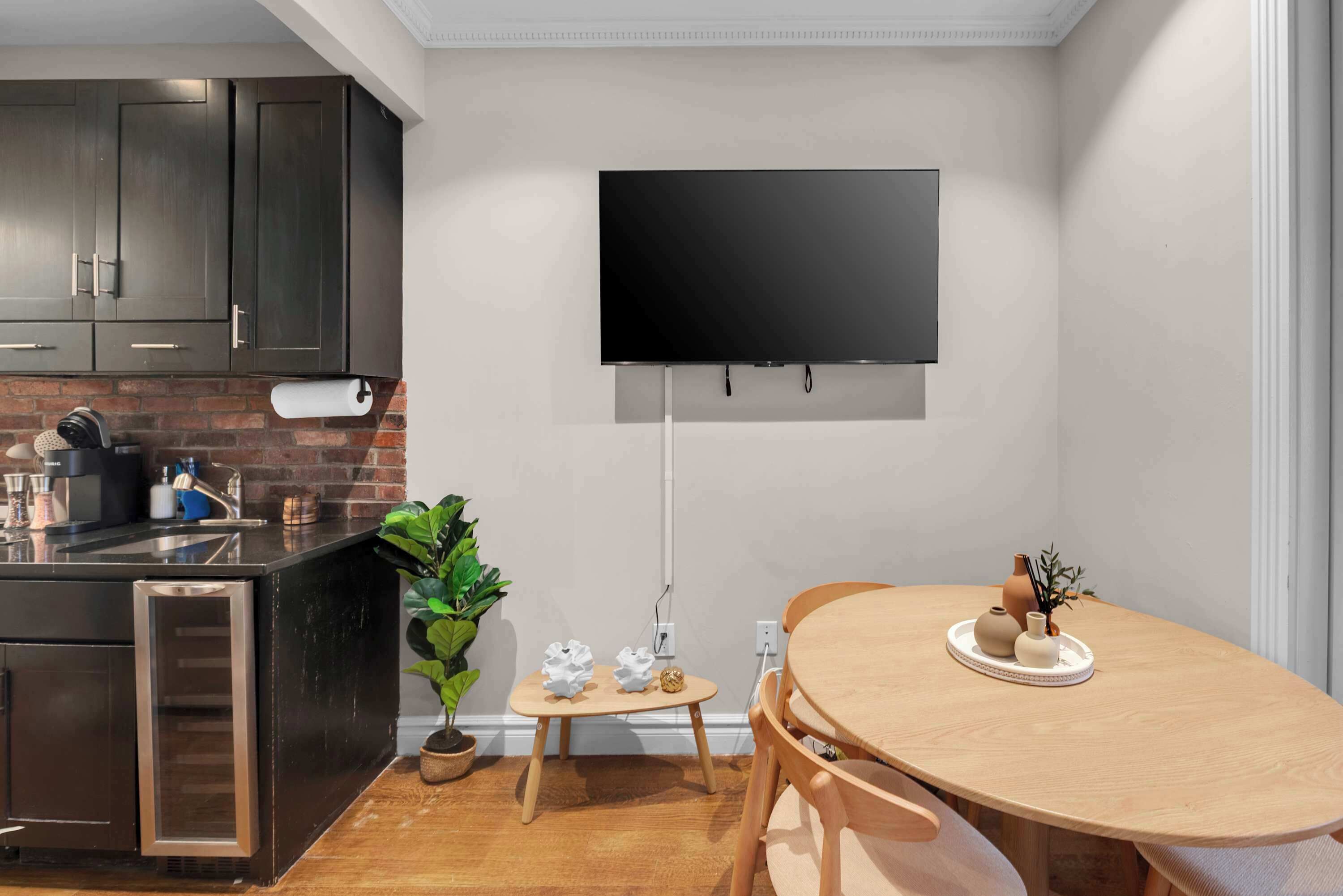 A modern kitchen with dark cabinets, a brick backsplash, a built-in refrigerator, a wooden dining table with chairs, a small side table, and a flat-screen TV mounted on the wall.