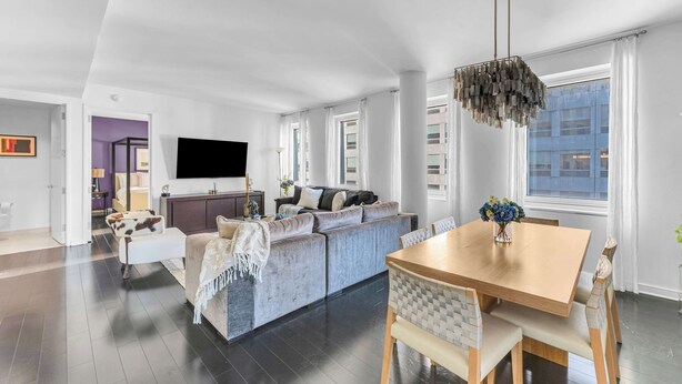 A modern living room with a grey sofa, a wooden dining table, and a TV mounted on the wall.