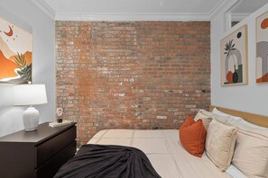 A bedroom with a brick wall, a bed with beige bedding, a nightstand with a lamp, and artwork on the walls. - Dream NYC Haven: Cozy 2BR in Prime Spot (New York)