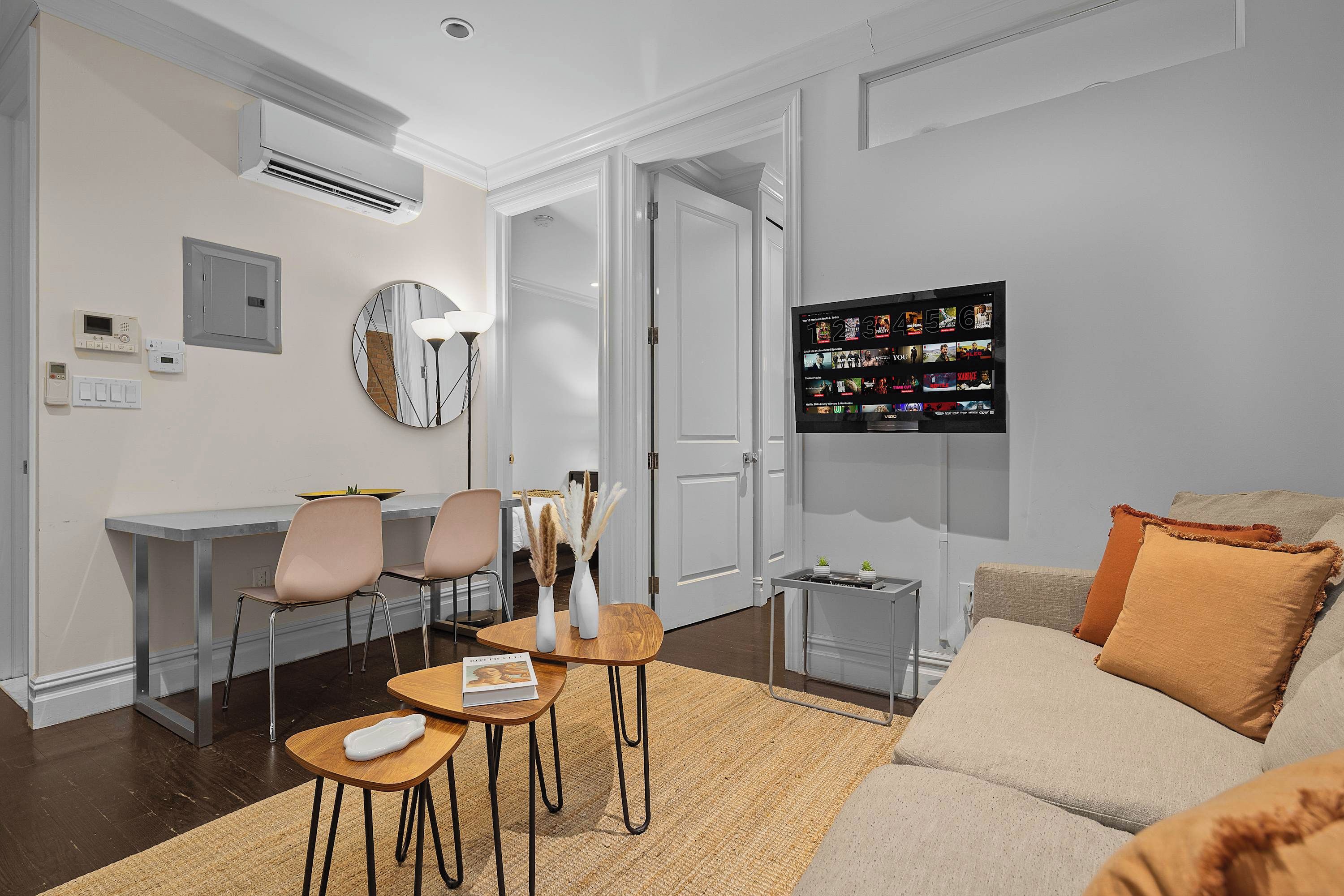 A modern living room with a sofa, a coffee table, and a dining area with chairs. There is a TV mounted on the wall and an air conditioning unit above the dining area.