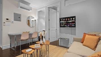 A modern living room with a sofa, a coffee table, and a dining area with chairs. There is a TV mounted on the wall and an air conditioning unit above the dining area.