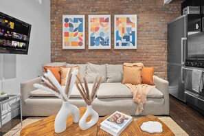 A modern living room with a brick wall, a flat-screen TV, a sofa with cushions, a coffee table with a book and decorative items, and a kitchen area with a refrigerator. - Dream NYC Haven: Cozy 2BR in Prime Spot (New York)