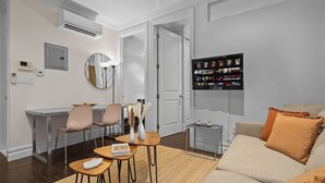 Ein modernes Wohnzimmer mit einer Couch, einem Couchtisch und einem Essbereich mit Stühlen. An der Wand ist ein Fernseher angebracht und über dem Essbereich befindet sich eine Klimaanlage.