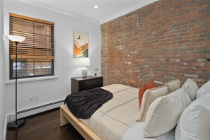 Un dormitorio con pared de ladrillo, cama con almohadas, mesita de noche con lámpara y ventana con persianas.