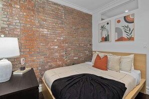 A hotel room with a brick wall, a bed with pillows, a bedside table with a lamp, and artwork on the wall. - Dream NYC Haven: Cozy 2BR in Prime Spot (New York)