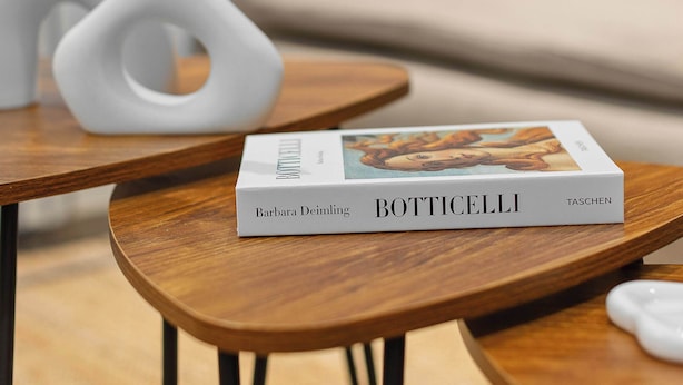 Ein Buch mit dem Titel 'Botticelli' auf einem Holztisch in einem Wohnzimmer im modernen Stil, inklusive einer weißen Vase mit Pampasgras und einem grauen Sofa mit orangefarbenen Kissen.