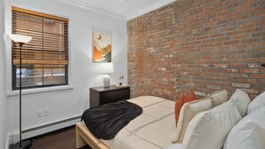 Ein Schlafzimmer mit Backsteinwand, einem Bett mit Kissen, einem Nachttisch mit Lampe und einem Fenster mit Jalousien.