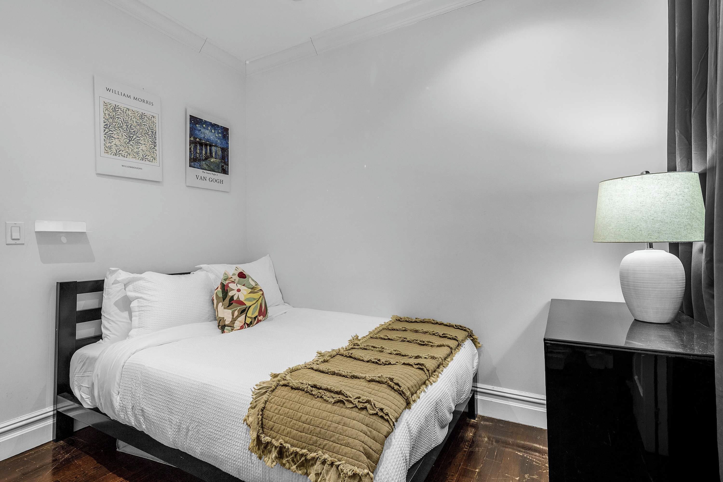 A bedroom with a bed, a bedside table with a lamp, and two framed artworks on the wall.