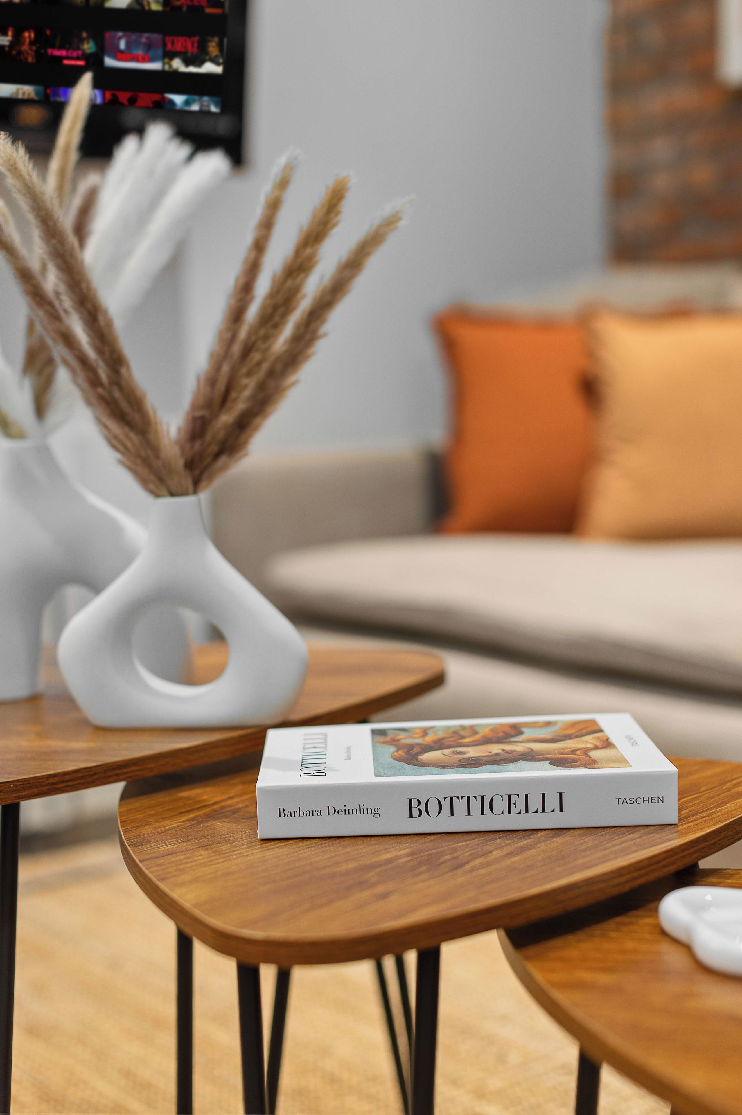 Un libro titulado 'Botticelli' sobre una mesa de madera en un salón con diseño moderno, que incluye un jarrón blanco con plumas de pampa y un sofá gris con cojines naranjas.