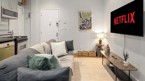 Una sala de estar moderna con un sofá gris, un televisor de pantalla plana y una zona de cocina con estufa y microondas.