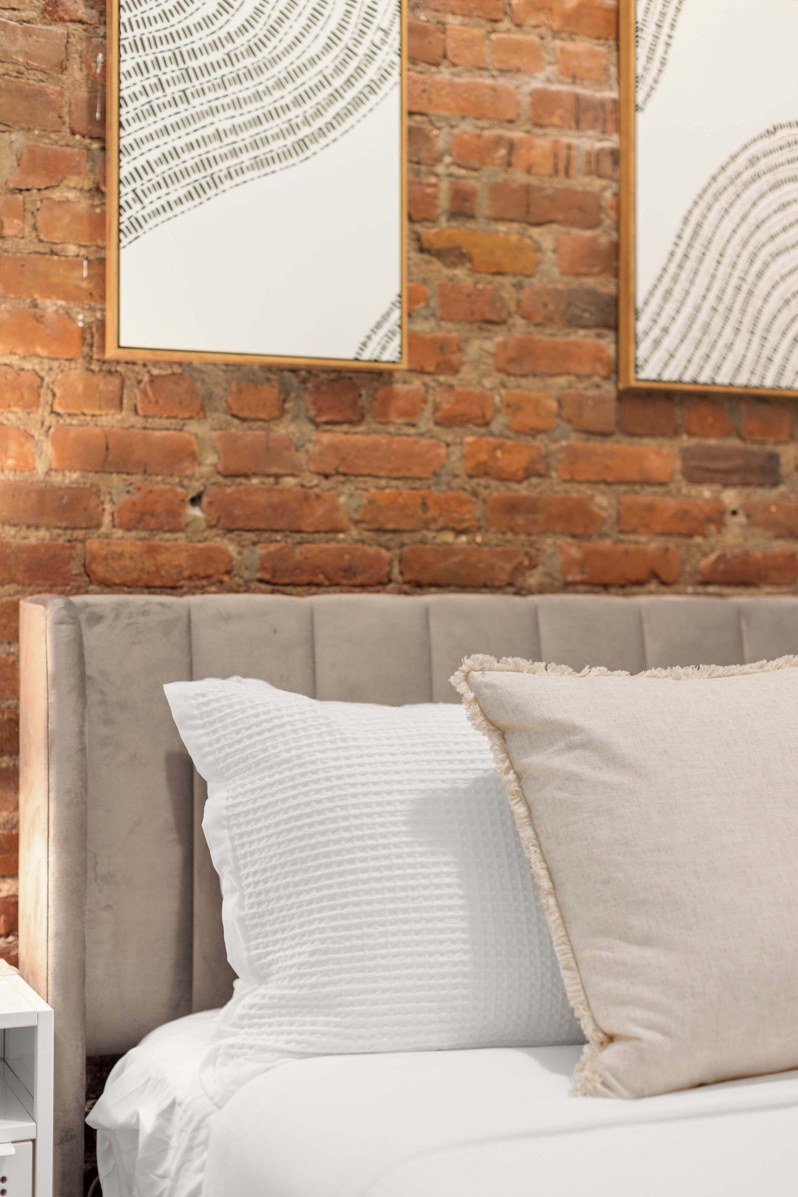Un lit avec une literie blanche et deux oreillers, une tête de lit rembourrée de couleur grise et un mur en briques orné de deux œuvres d’art abstraites encadrées.