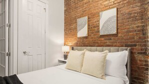 Un dormitorio con pared de ladrillo, una puerta blanca, una cama con ropa de cama blanca y dos cuadros enmarcados.