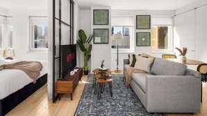 Un salon moderne avec un canapé gris, une table basse et un téléviseur. Une grande fenêtre offre une vue sur les bâtiments à l’extérieur.