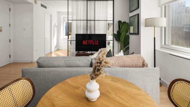 Un salon moderne avec un canapé gris, une table basse en bois sur laquelle est posé un vase blanc, et un téléviseur qui affiche le logo « NETFLIX » en arrière-plan.