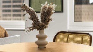 Une table ronde en bois surmontée d’un vase blanc contenant des plantes séchées, et flanquée de deux chaises en osier avec des coussins verts.