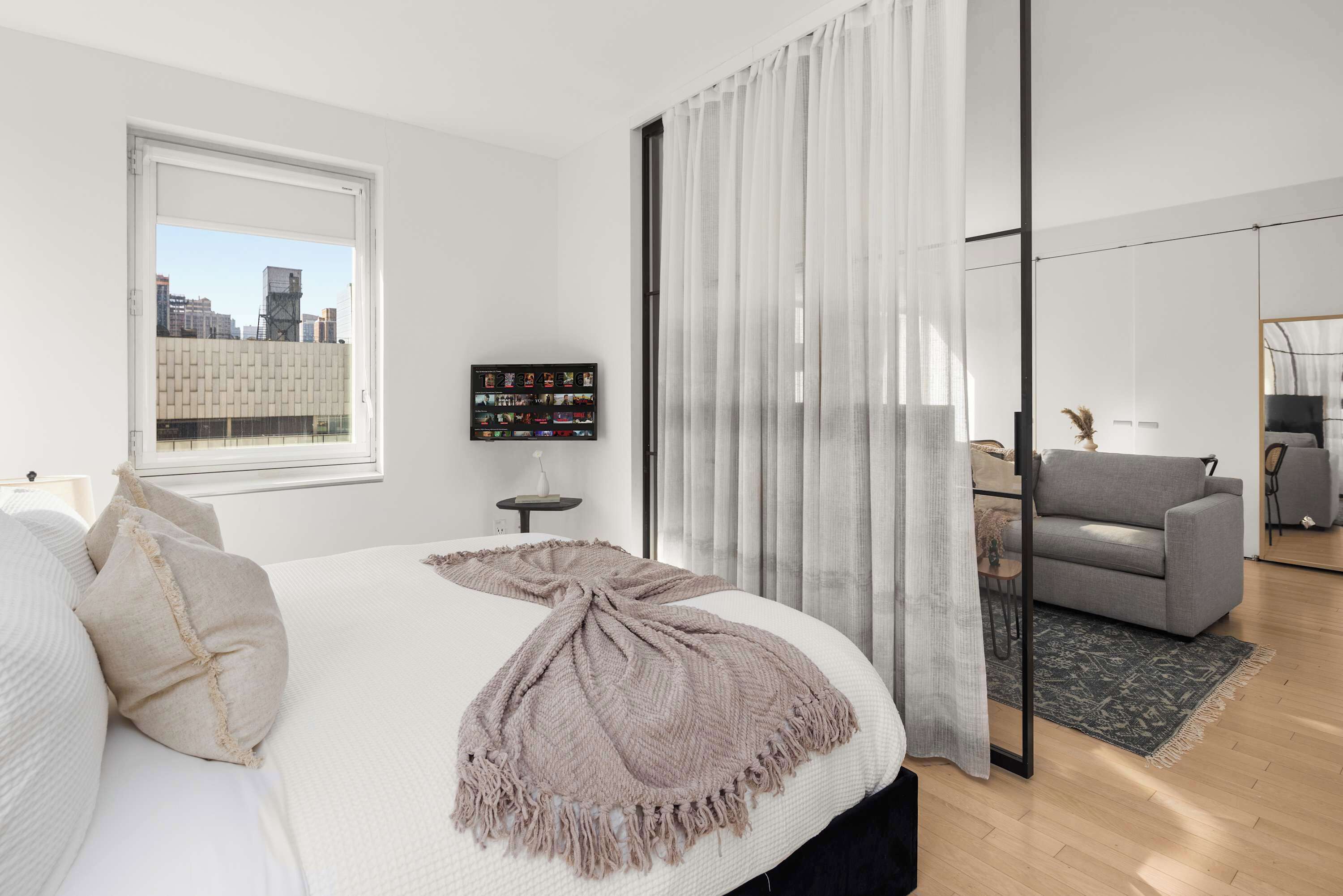 Een moderne slaapkamer met een groot bed, uitzicht op de stad, een grijze bank en een klein tafeltje met een decoratief object.