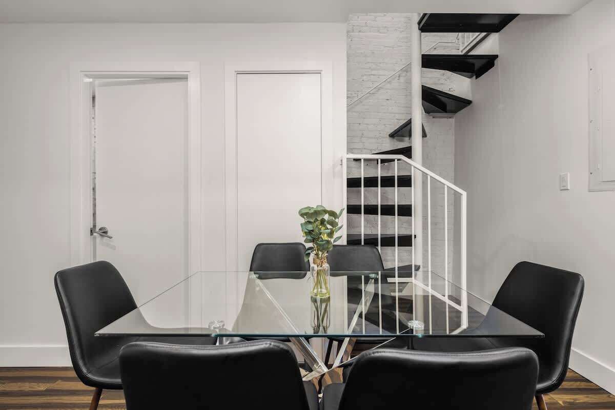 Un espace repas moderne avec une table en verre et des chaises noires, une porte blanche et un escalier avec un mur blanc et des rampes noires.