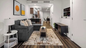 Uma sala de estar moderna com um sofá cinza, uma mesa de centro e um tapete. Cozinha visível ao fundo, com um espaço de refeições.