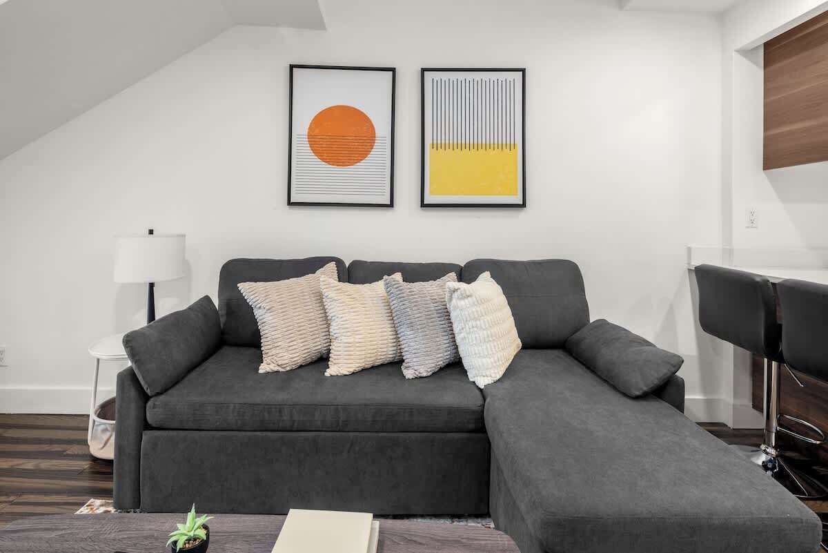 Een moderne woonkamer met een donkergrijze hoekbank, meerdere kussens, een kleine potplant en twee kunstwerken in lijsten aan de muur.