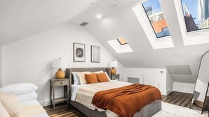 Quarto moderno com teto inclinado, claraboias, cama com manta marrom, mesa de cabeceira com abajur e um espelho grande.