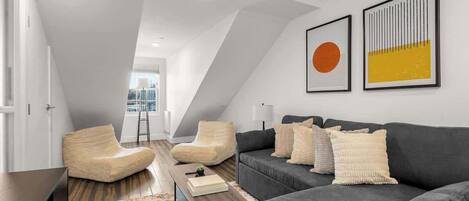 Um quarto moderno no estilo loft, com teto inclinado, um sofá cinza escuro, duas poltronas e uma mesa de centro de madeira.