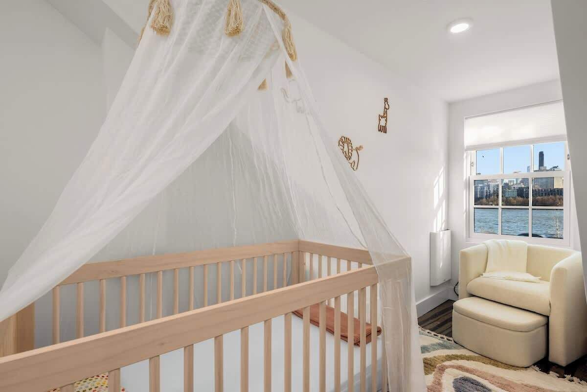 Una habitación con una cuna de madera, un dosel blanco, vistas a la ciudad a través de una ventana y un sillón color beige.