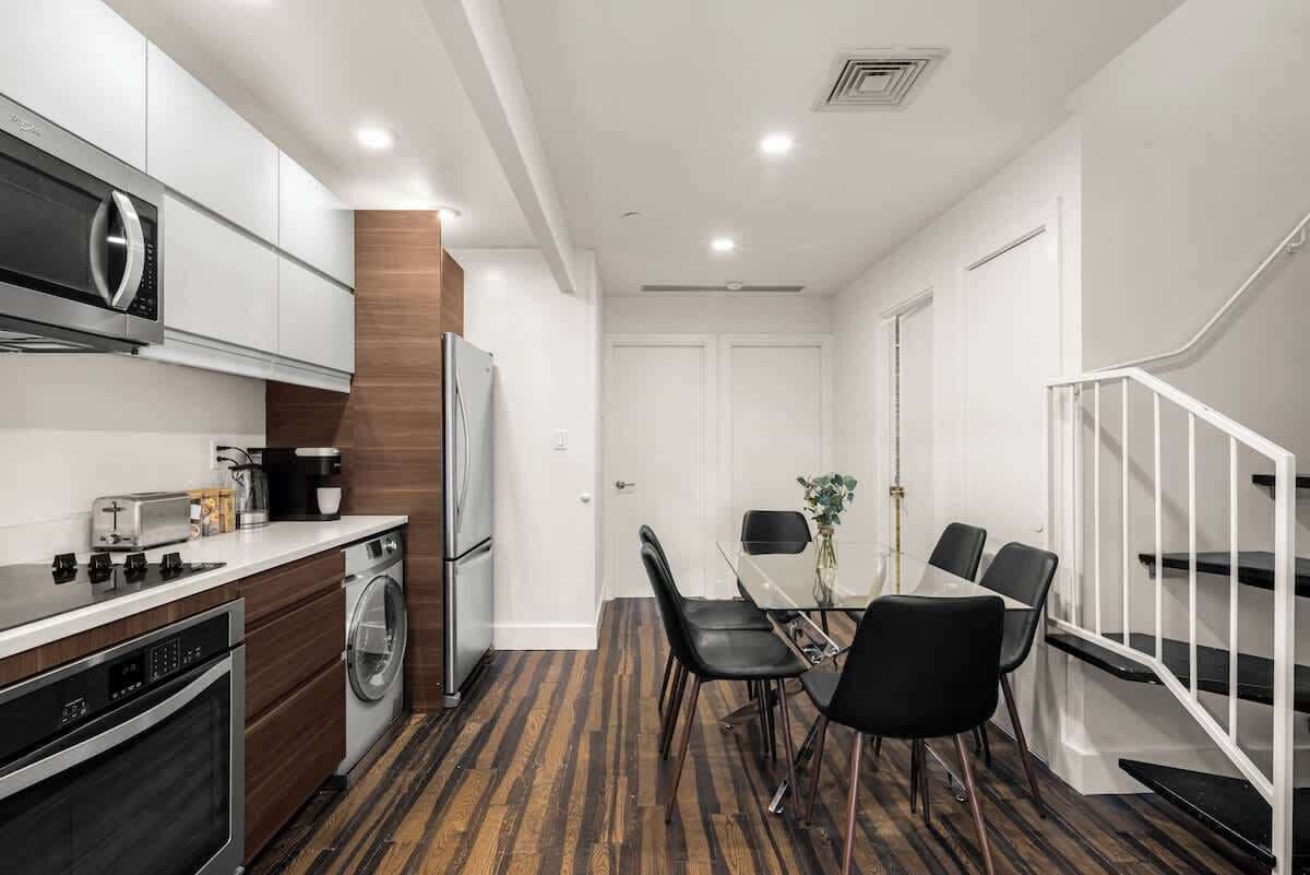 Una cocina moderna con electrodomésticos de acero inoxidable, un comedor con una mesa de vidrio y sillas negras, y una escalera con barandillas blancas.