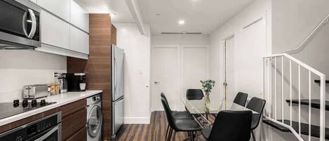 A modern kitchen with stainless steel appliances, a dining area with a glass table and black chairs, and a staircase with white railings.
