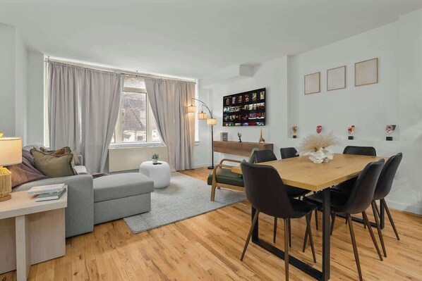 A modern living room with a sofa, a dining table with chairs, a coffee table, and a TV mounted on the wall. - Spacious 2BR Steps from Time Square & Hudson Yards (New York)