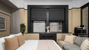 A bedroom with a bed, a grey sofa, a window with blinds, a plant, and a decorative wall piece. - Central Midtown 1BR | Walk to Times Square! (New York)