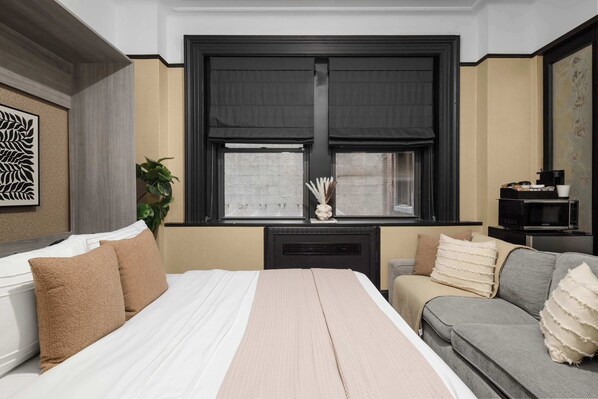 A bedroom with a bed, a grey sofa, a window with blinds, a plant, and a decorative wall piece.