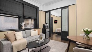 A modern living room with a grey sofa, a black coffee table, and a dining area with a wooden table and chairs. - Central Midtown 1BR | Walk to Times Square! (New York)