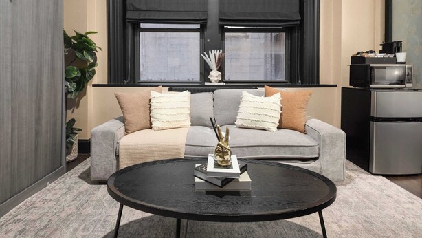 A modern living room with a grey sofa, two cushions, a black coffee table, and a small decorative item. - Central Midtown 1BR | Walk to Times Square! (New York)