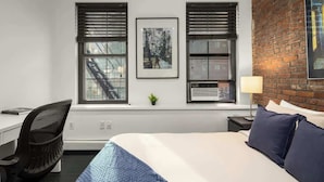 Ein Hotelzimmer mit Bett, Schreibtisch, zwei Fenstern mit Jalousien, einem gerahmten Bild an der Wand und einer Wand aus Ziegelsteinen.