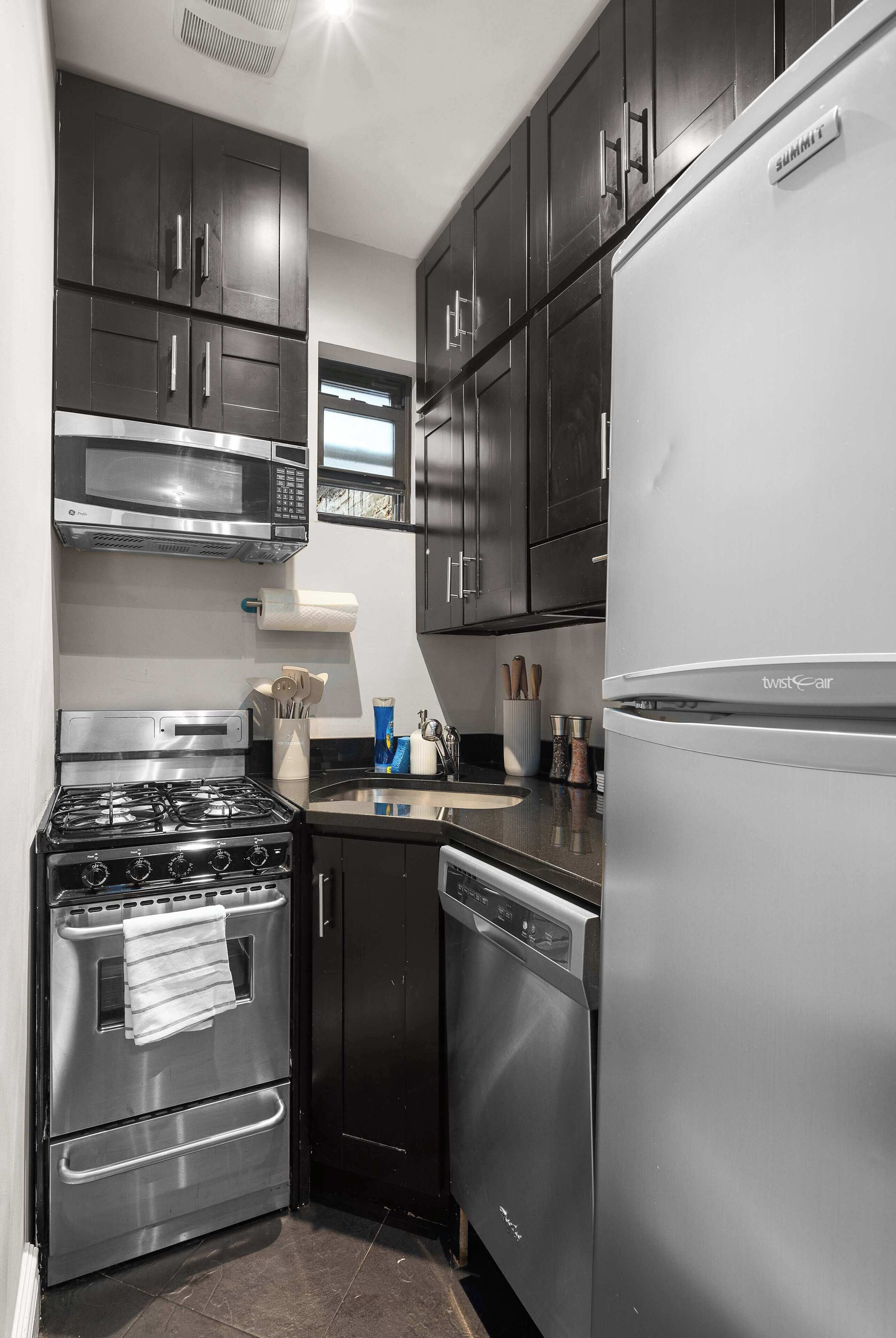 Una cocina compacta con armarios oscuros, electrodomésticos de acero inoxidable, incluyendo estufa, microondas y lavavajillas, además de un refrigerador.