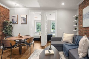 A modern living room with a dining area, a grey sofa, and a brick accent wall. - Stylish Retreat in Midtown NYC Walk to Everything (New York)