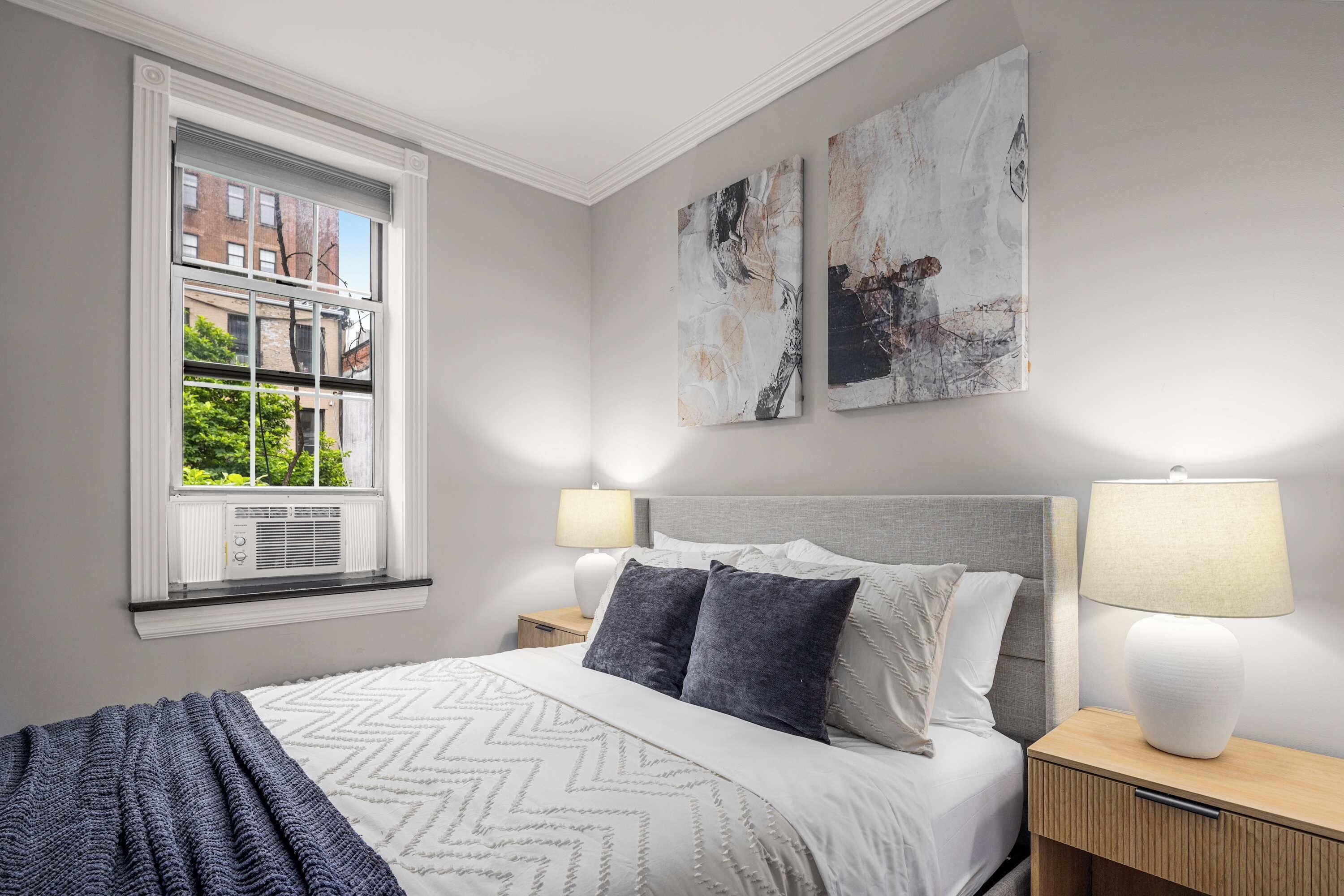 Ein ordentlich eingerichtetes Schlafzimmer mit einem großen Bett, zwei Nachttischen und einem Fenster mit Blick auf Gebäude und Bäume.