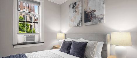 Ein ordentlich eingerichtetes Schlafzimmer mit einem großen Bett, zwei Nachttischen und einem Fenster mit Blick auf Gebäude und Bäume.