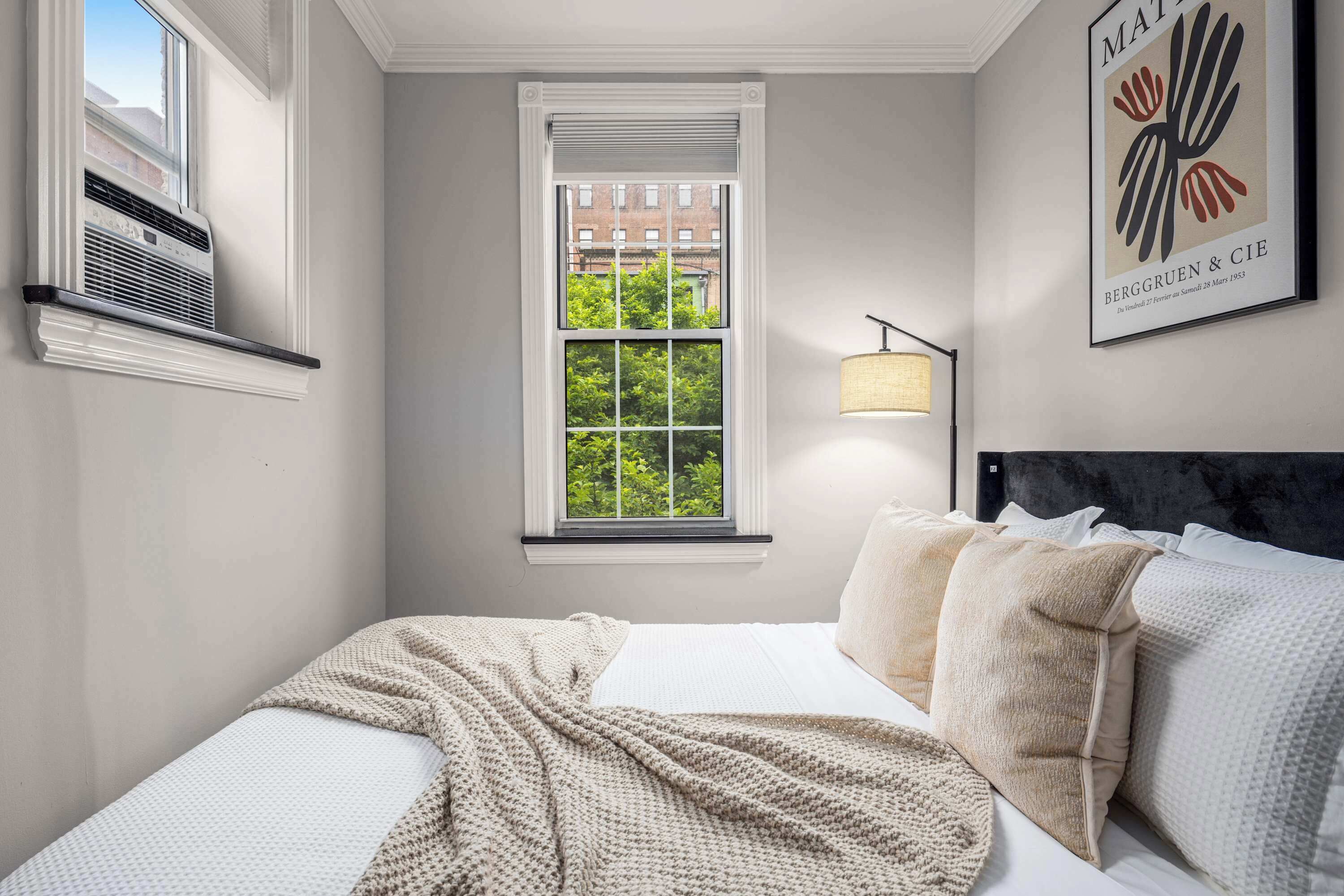 A bedroom with a large window, a bed with multiple pillows, a wall-mounted air conditioner, and a framed artwork.