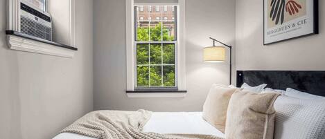 A bedroom with a large window, a bed with multiple pillows, a wall-mounted air conditioner, and a framed artwork.