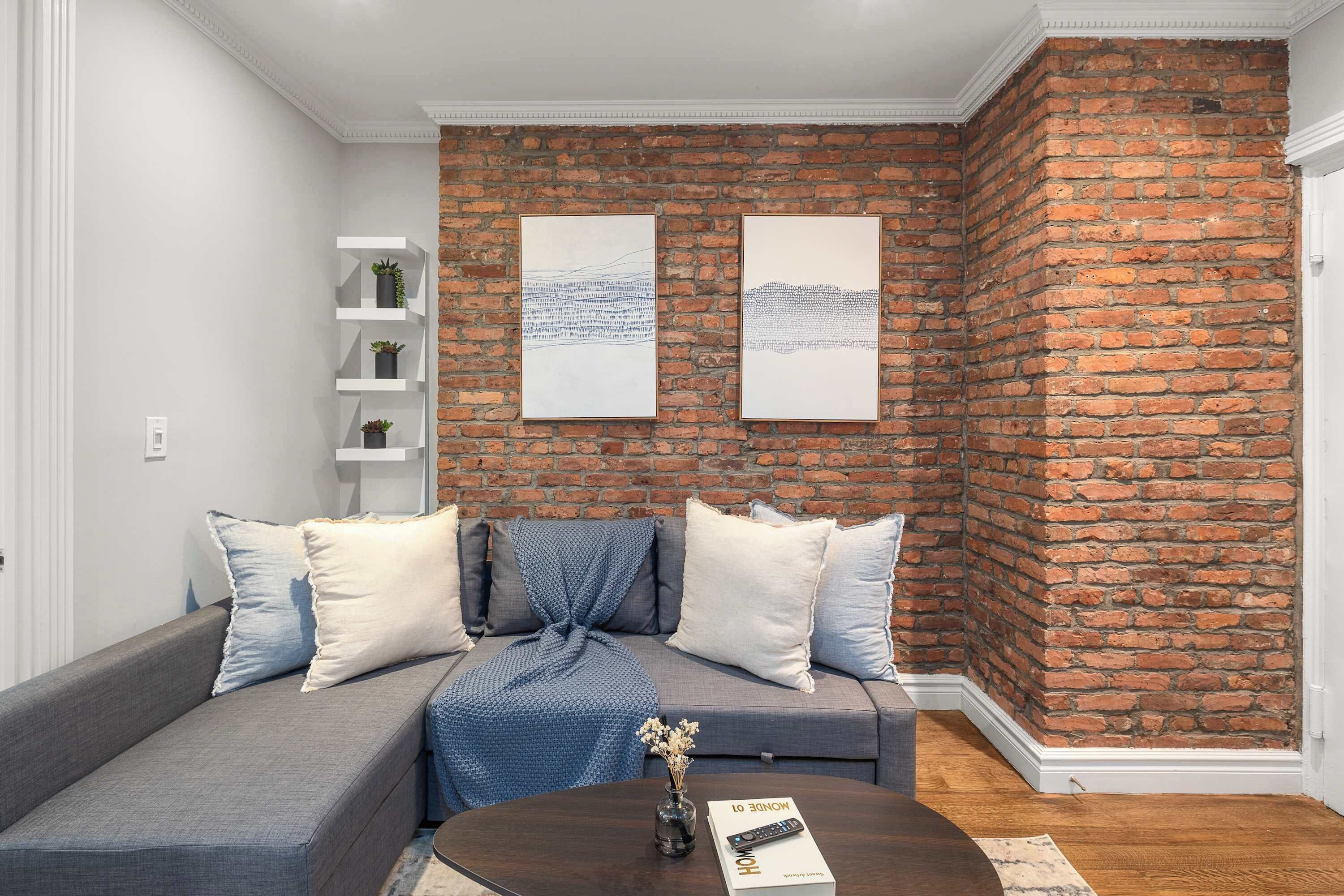 Een moderne woonkamer met een accentmuur van baksteen, een grijze bank met witte en blauwe kussens, en een kleine tafel met een vaas en een afstandsbediening.