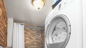 Une petite buanderie comprenant une machine à laver et un sèche-linge superposés, un espace douche avec un mur en briques et des toilettes.