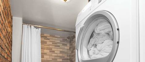 A compact laundry room with a stacked washer and dryer, a shower area with a brick wall, and a toilet.