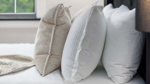 Un lit bien fait, avec une literie blanche et plusieurs oreillers, une lampe sur pied et une fenêtre donnant sur un paysage verdoyant.