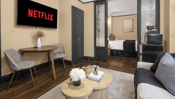 A modern hotel room with a flat-screen TV displaying "NETFLIX," a round wooden coffee table with a vase and book, a gray armchair, a sofa, and a bed with a black headboard. - Premier Midtown Stay | Walk to Times Square! (New York)