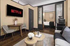 A modern hotel room with a flat-screen TV displaying "NETFLIX," a round wooden coffee table with a vase and book, a gray armchair, a sofa, and a bed with a black headboard.